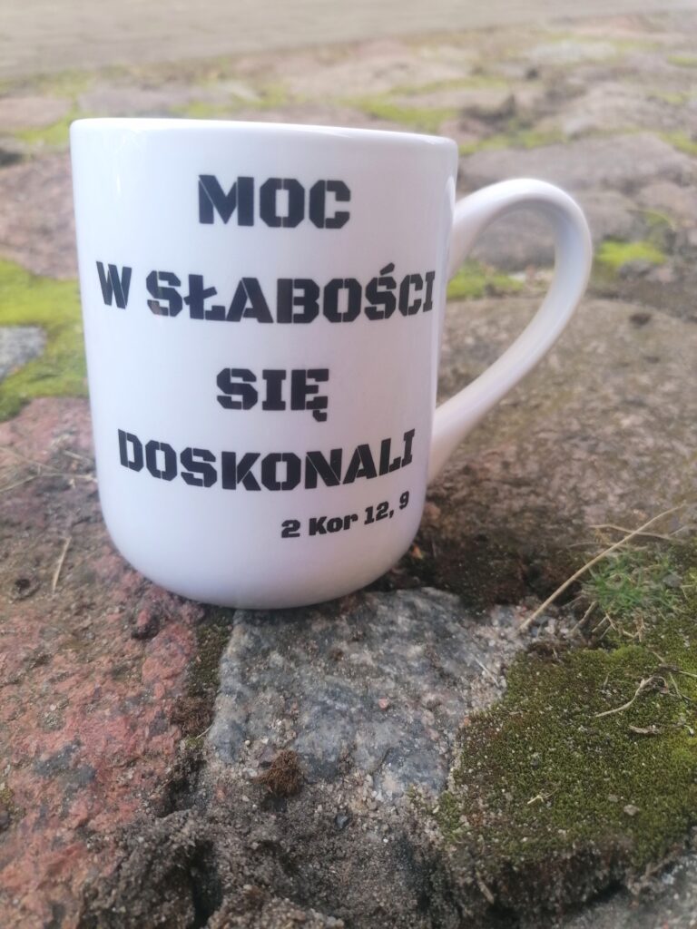 Biały kubek ceramiczny z militarnym napisem Moc w słabości się doskonali 2 Kor 12 9 stojący na kamiennym podłożu