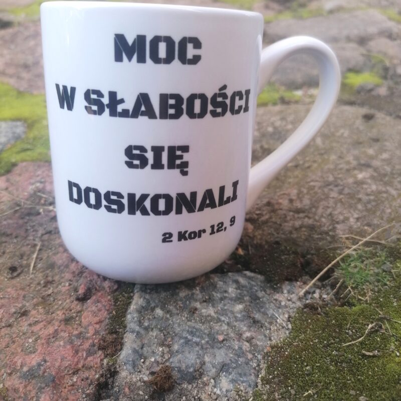 Biały kubek ceramiczny z militarnym napisem Moc w słabości się doskonali 2 Kor 12 9 stojący na kamiennym podłożu