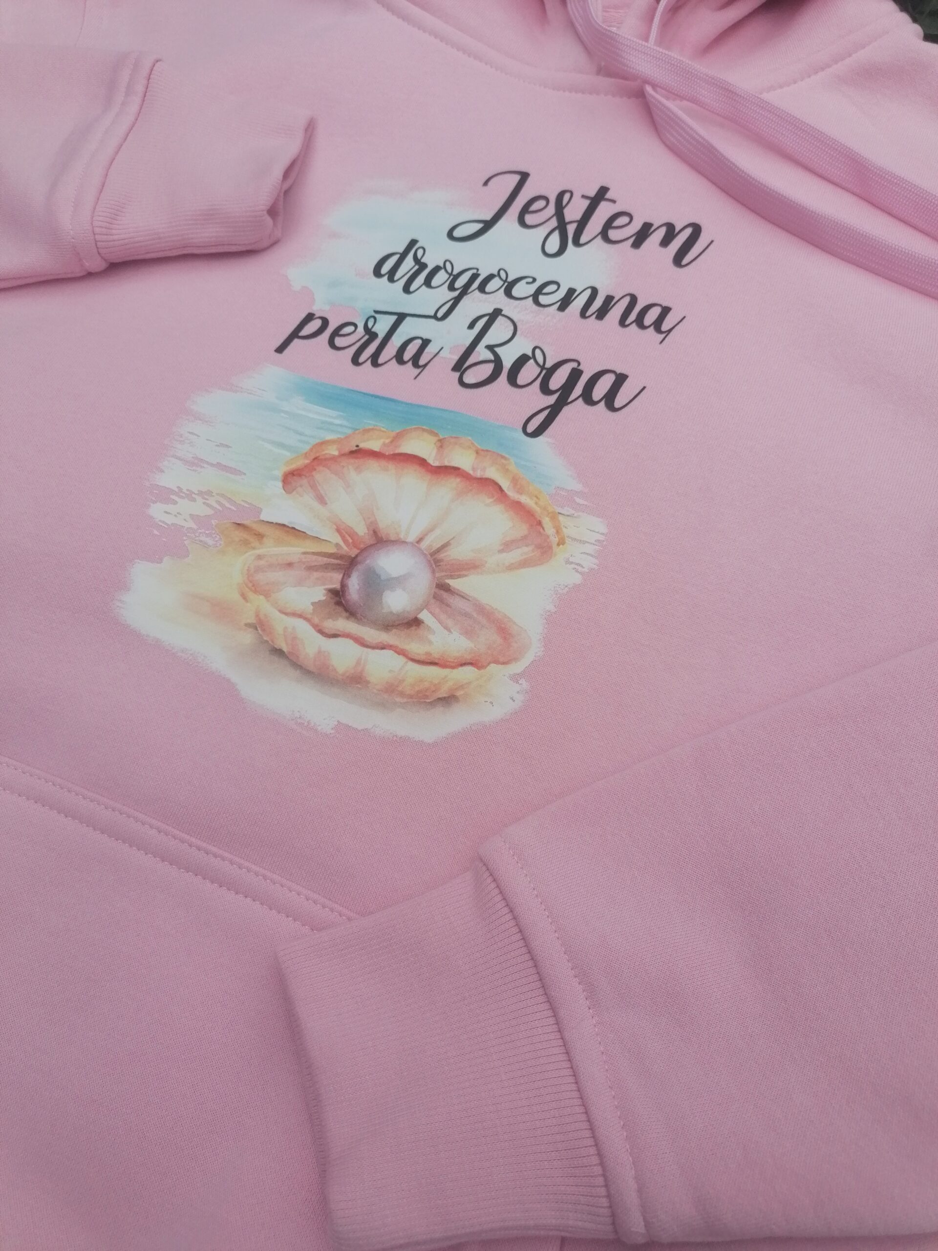 Bluza z kapturem „Jestem drogocenną perłą Boga” 2 Zbliżenie napisu „Jestem drogocenną perłą Boga” i grafiki muszli z perłą na różowej bluzie.