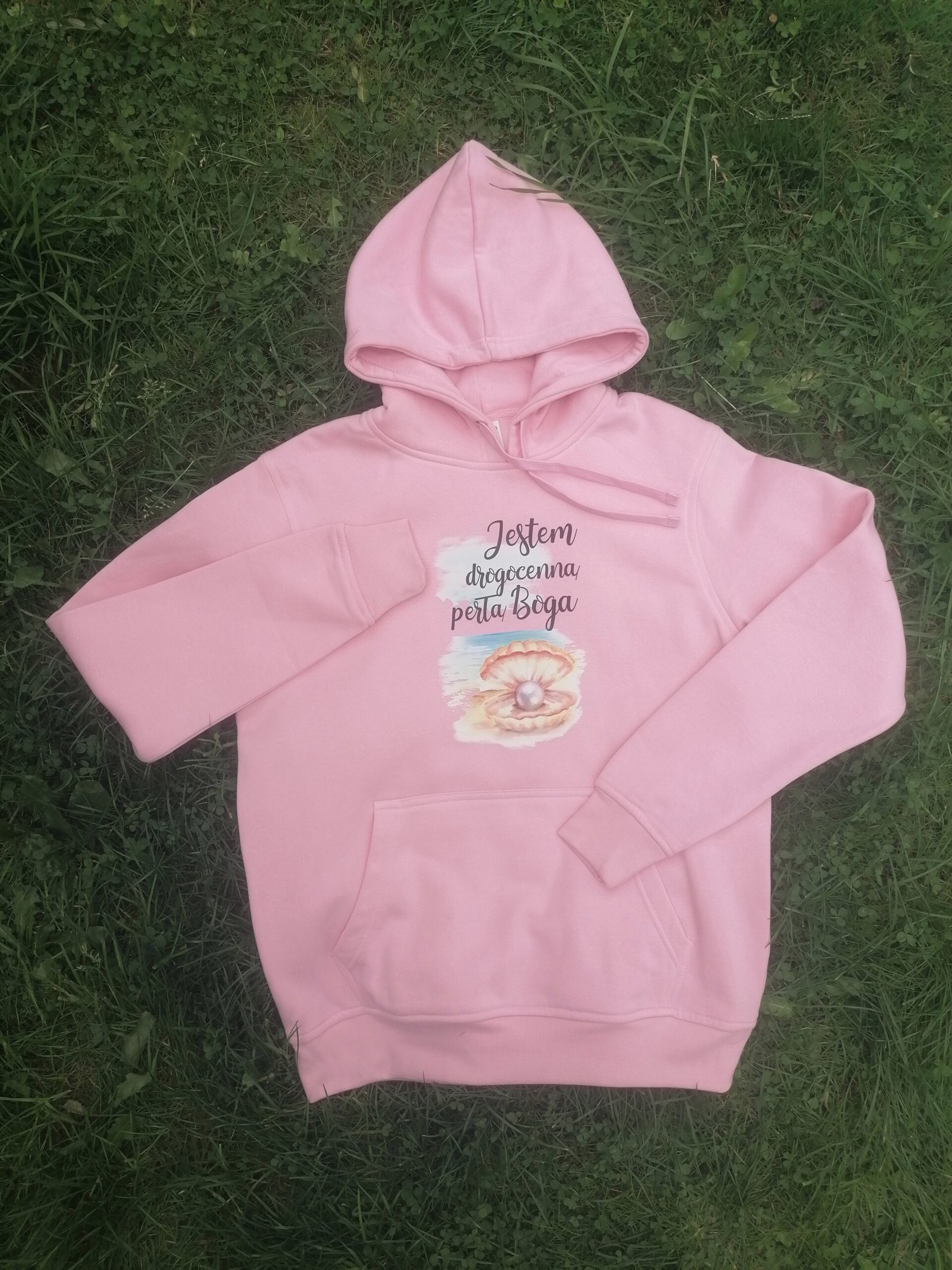 Bluza z kapturem „Jestem drogocenną perłą Boga” 3 Różowa bluza z kapturem „Jestem drogocenną perłą Boga” i kieszenią kangurką, ułożona na trawie.